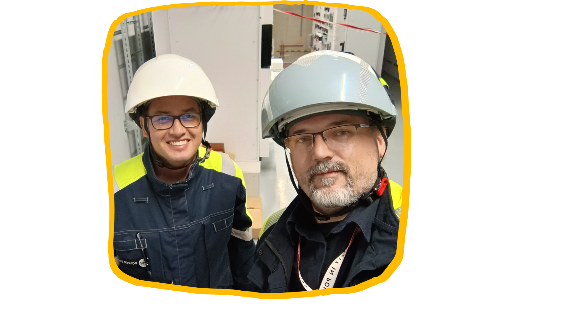 Two workers wearing safety helmets and protective gear, smiling while standing in an industrial setting with equipment and safety barriers in the background.